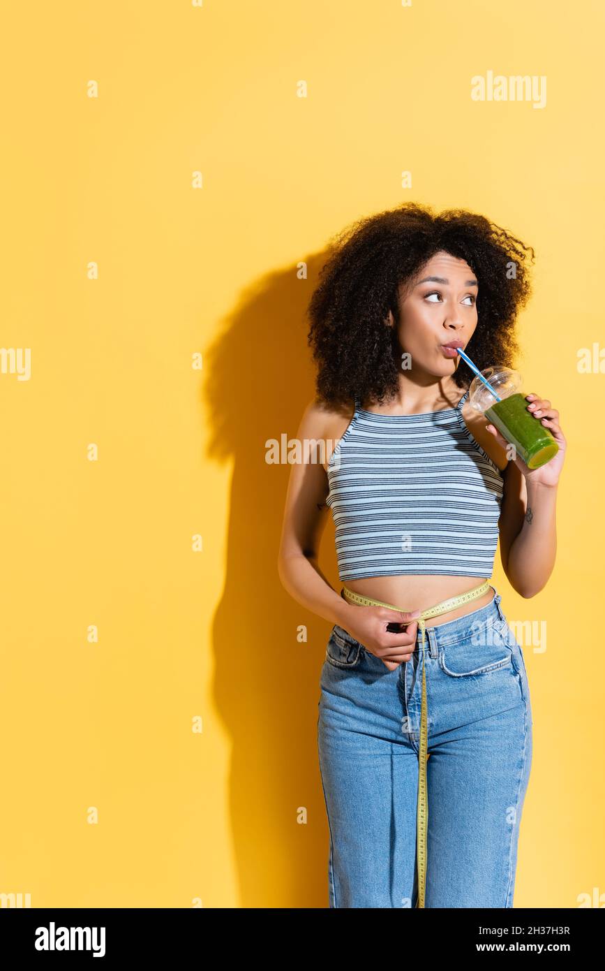 mujer afroamericana que mide la cintura mientras bebe batido fresco en amarillo Foto de stock