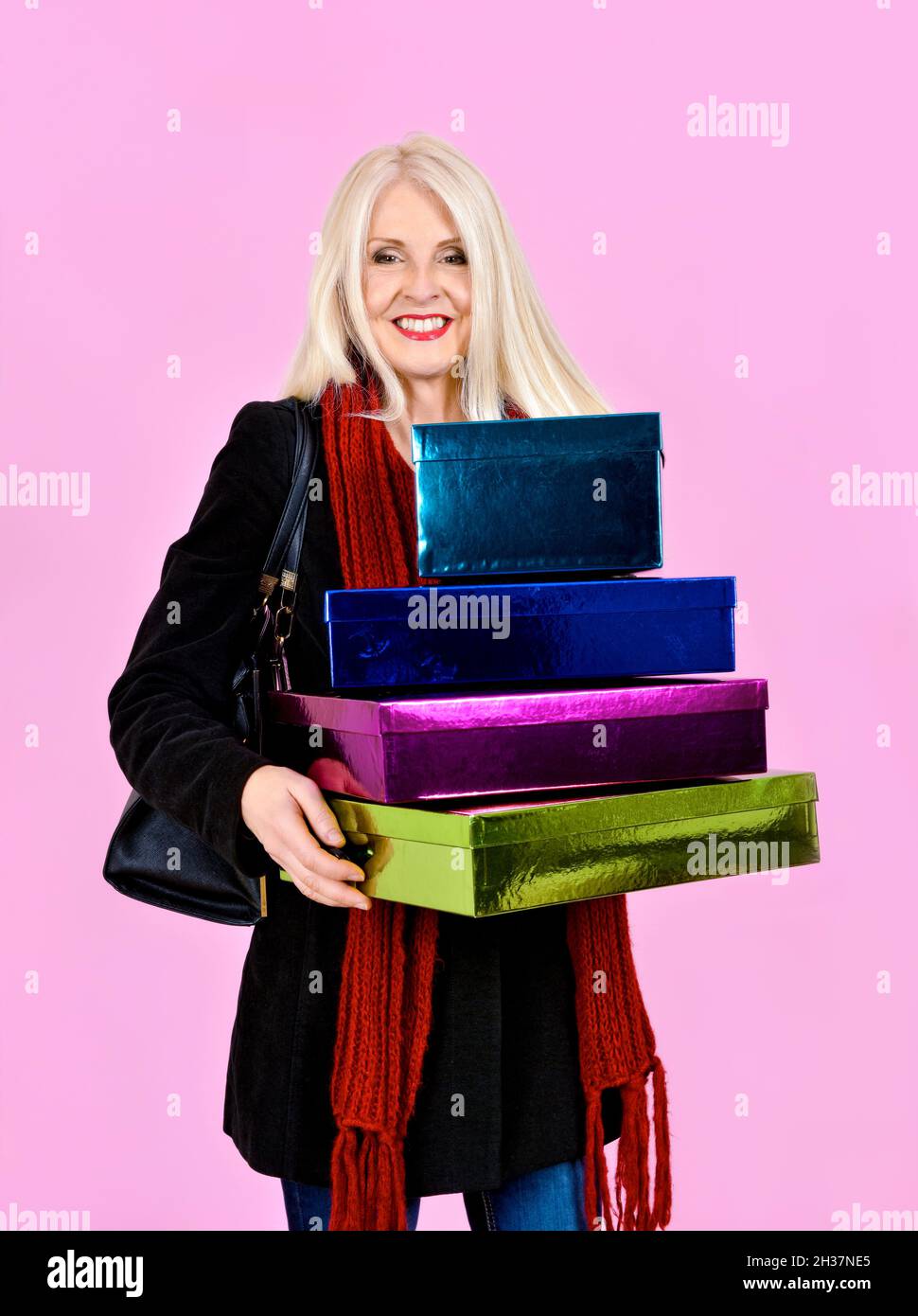 Atractiva dama rubia vestida de abrigo y bufanda riendo mientras llevaba un montón de cajas de colores o regalos, tomadas sobre fondo rosa Foto de stock