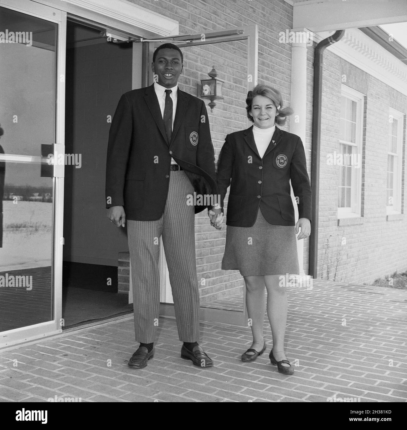 1967, histórico, un hombre negro sudente y una mujer blanca estudiante que lleva sus blazers de la universidad con logotipos, sosteniendo las manos con los otros en la puerta a las aulas, Virginia, EE.UU.. Son miembros del Consejo Estudiantil de la universidad. En esta era, el uso de blazers, tanto para niñas como para niños era común en la escuela secundaria y la universidad, ya que la educación era todavía un mundo razonablemente formal y tradicional. Se espera que los Boyswere usen camisas blancas con corbata debajo del blazer y las muchachas, un blazer y falda. Foto de stock