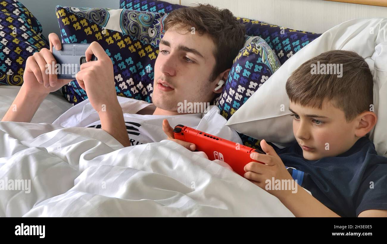 dos hermanos están acostados uno al lado del otro en la cama y enloquándose con un smartphone y una consola de juegos Foto de stock