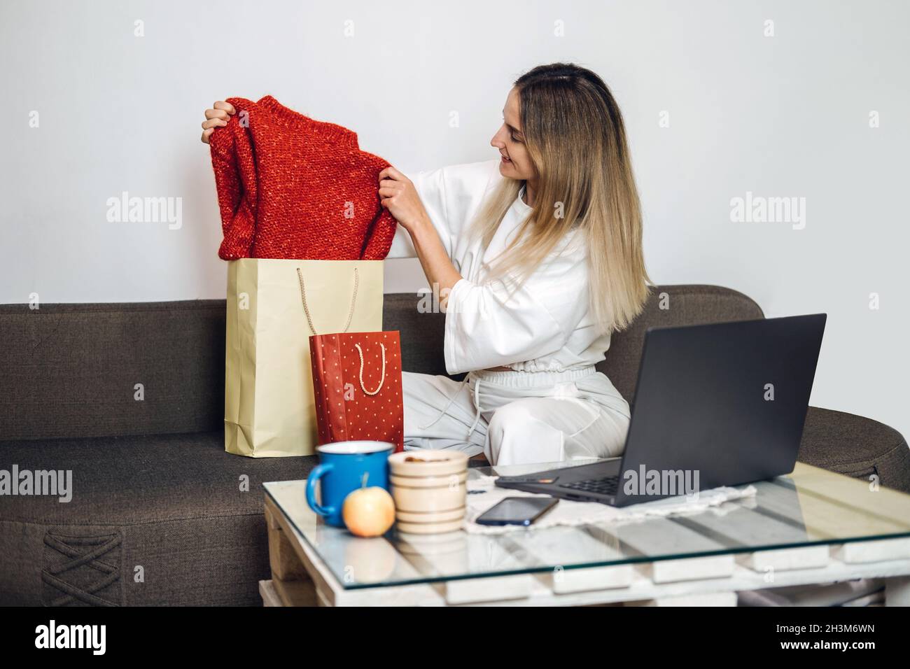 black friday, venta en línea, compras en Internet, comercio electrónico. La  mujer caucásica saca ropa nueva de la bolsa de la compra sobre el fondo del  portátil en casa. Mujer Fotografía de