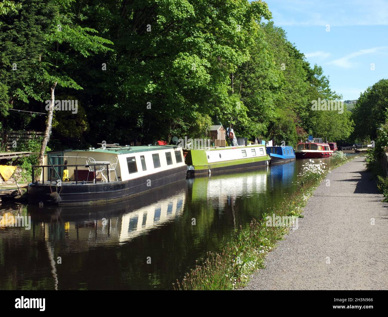 Barcos del canal amarrados frente al camino en el canal rochdale cerca del puente hebden rodeado de árboles bajo la luz del sol en verano Foto de stock