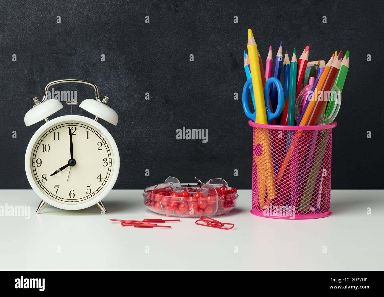 Reloj de alarma redondo blanco y cristal metálico con bolígrafos, lápices y rotuladores sobre el fondo de una pizarra negra vacía Foto de stock