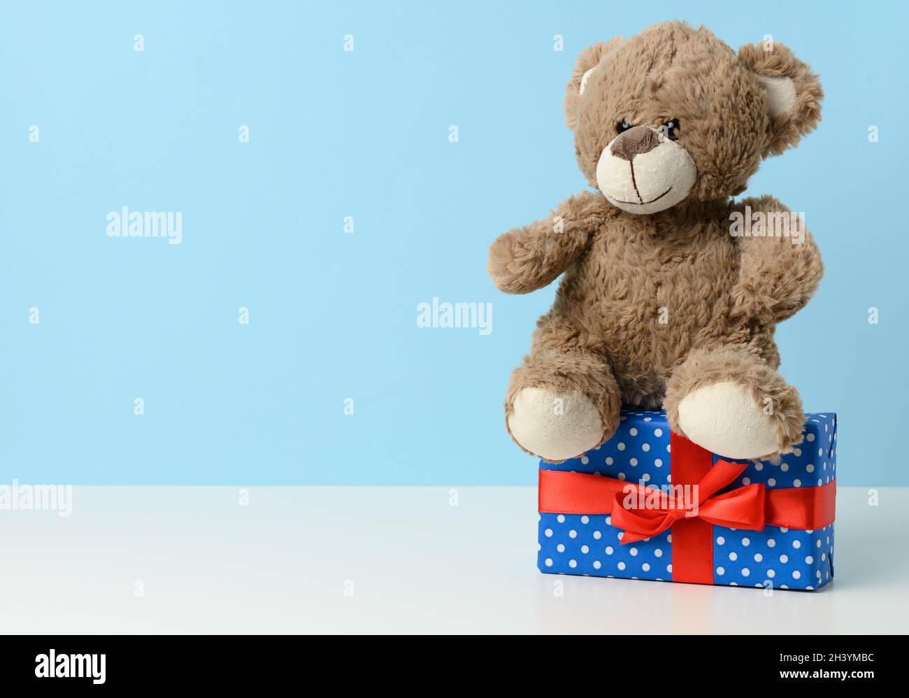 Lindo oso de peluche marrón sosteniendo una caja envuelta en papel azul y cinta de seda roja sobre mesa blanca. Premio y felicitaciones Foto de stock