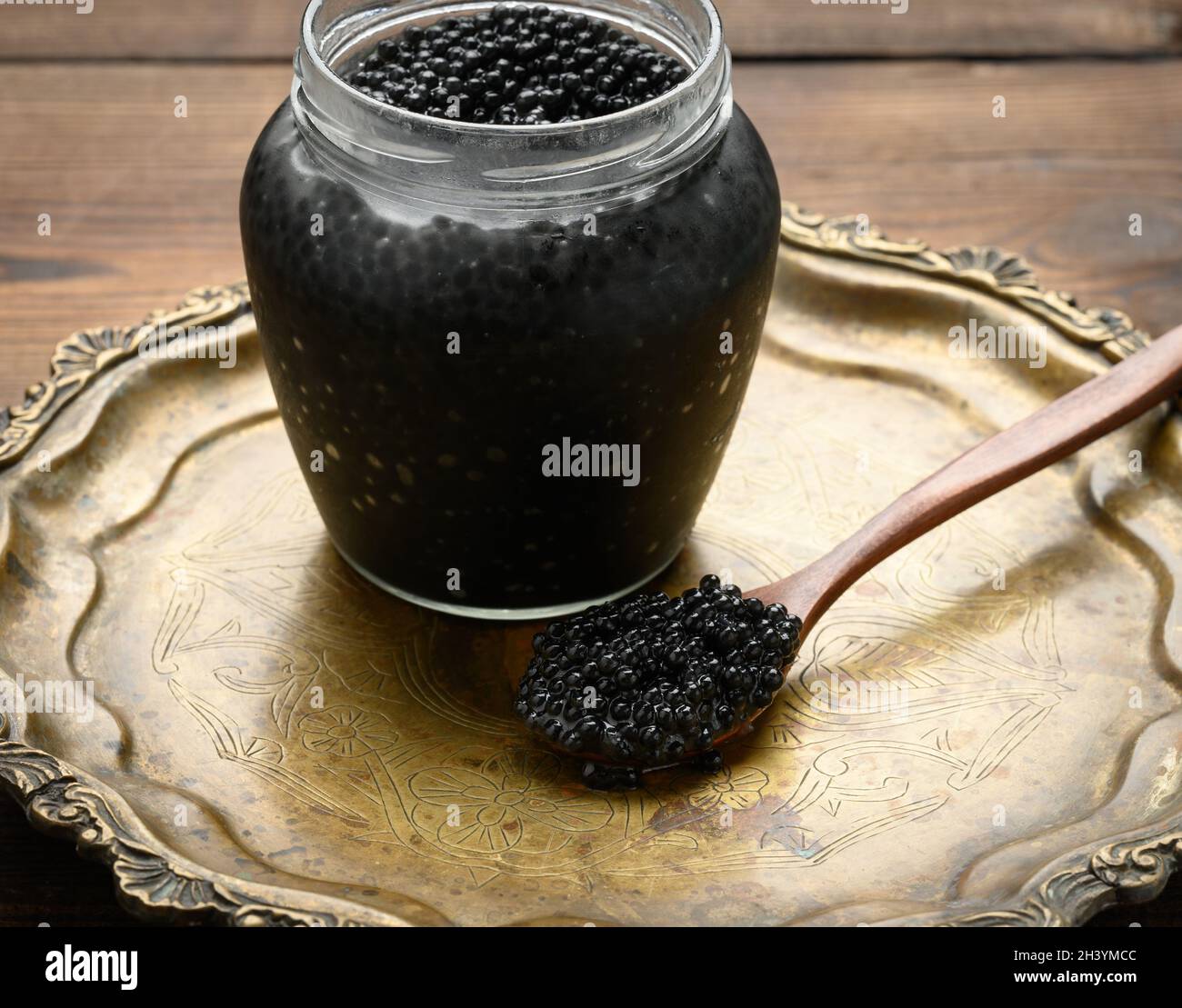 Caviar de pez espada negro fresco en una cuchara marrón de madera y una lata completa de caviar en una mesa de madera Foto de stock