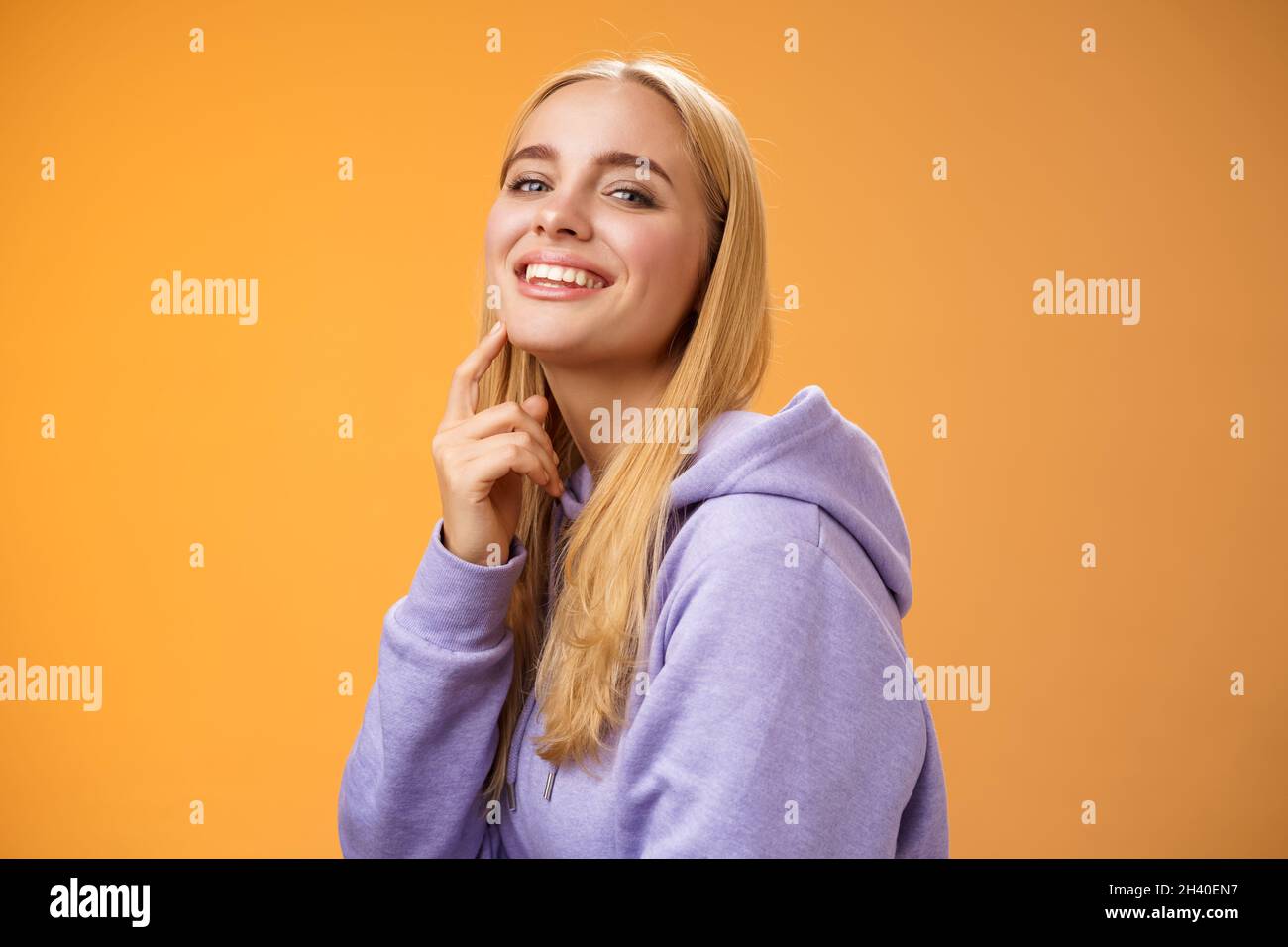 Atractiva y segura mujer blanca rubia en púrpura hoodie cámara de giro tocando la barbilla sonrisa cheeky cámara comprobar hacia fuera Foto de stock