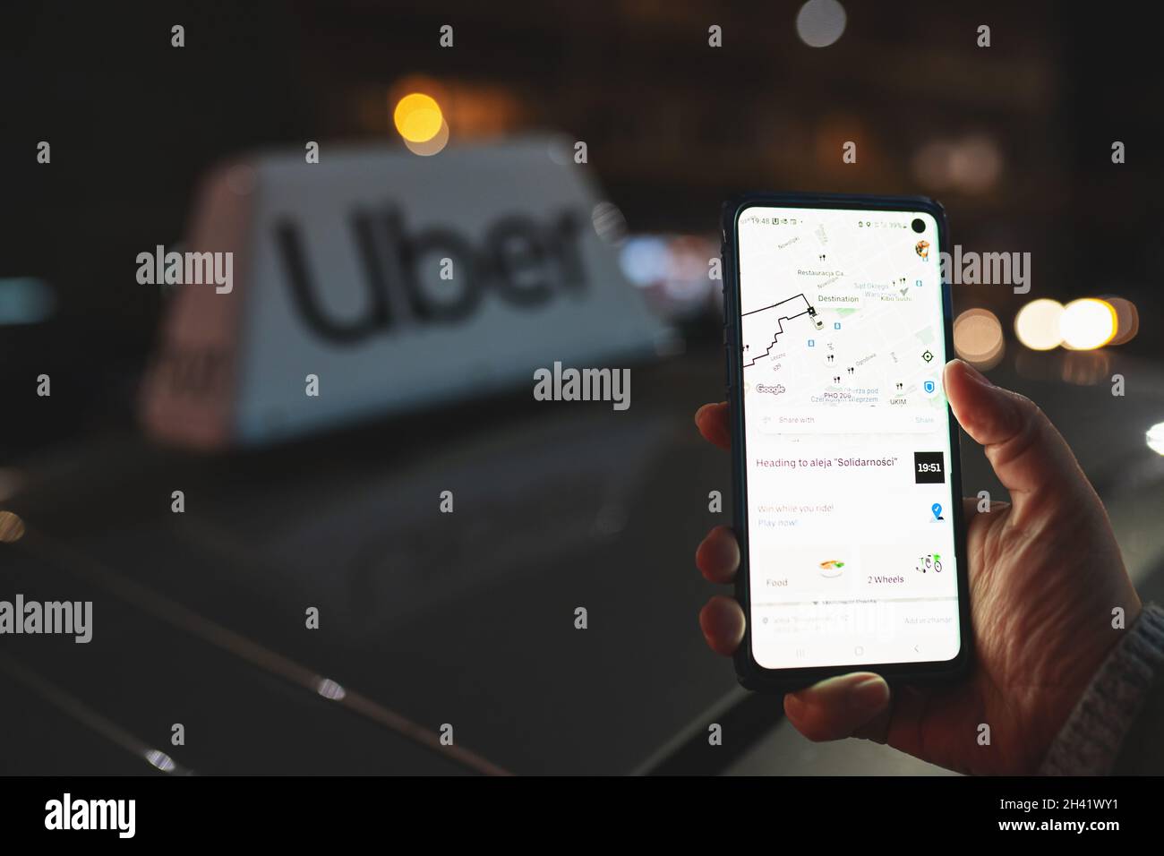 La aplicación Uber se muestra en el teléfono inteligente que se mantiene en la mano delante de la señal de taxi Uber en la parte superior de un coche por la noche con luces de la ciudad de fondo con enfoque suave. Un concepto de cabina. Varsovia, Polonia - 23 de octubre de 2021 Foto de stock