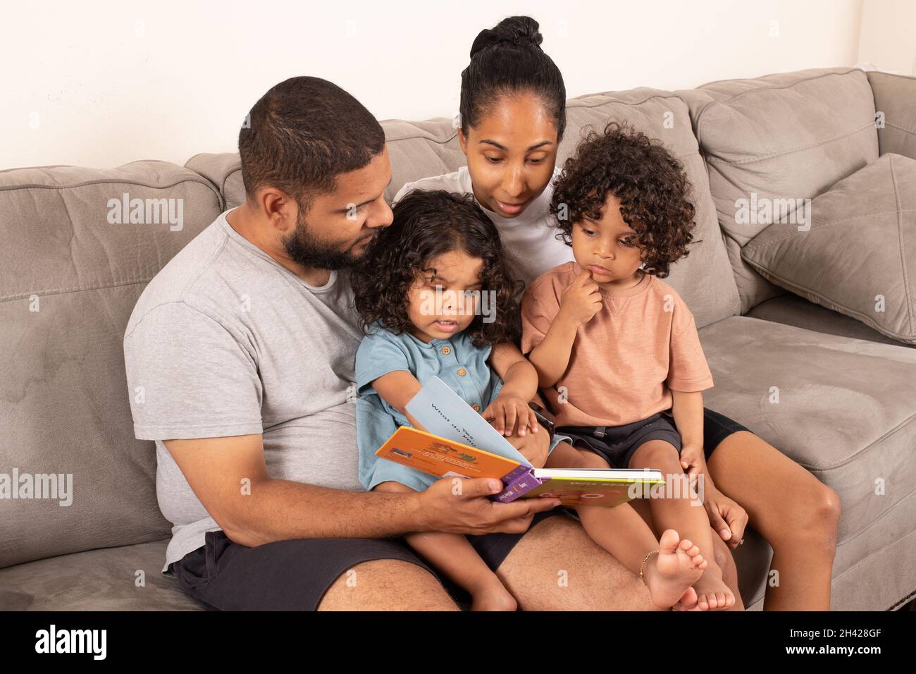 gemelos fraternos de 2 años, niño y niña, sentados en el sofá con los padres, leen a los libros de la mesa Foto de stock