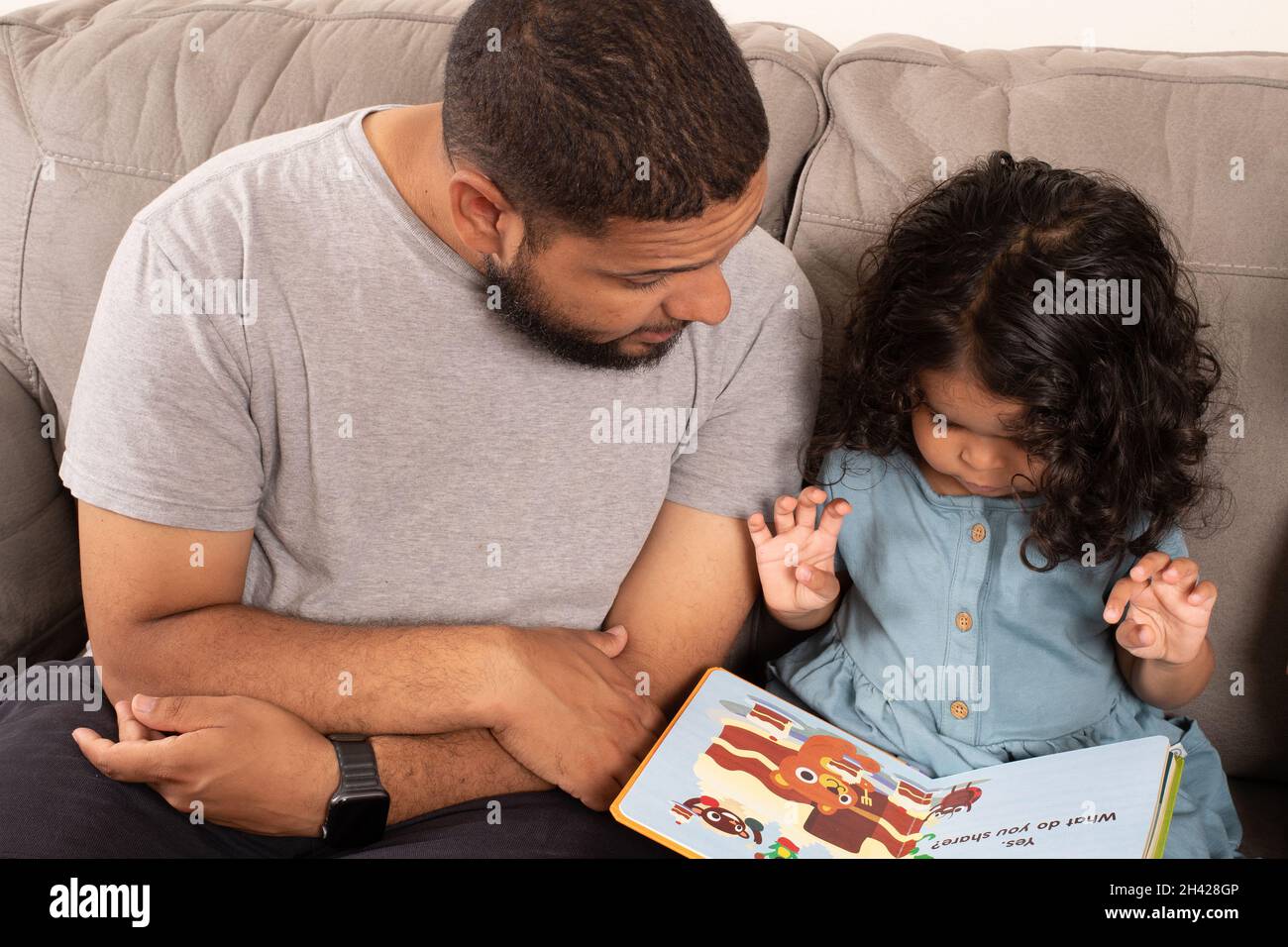 una niña de 2 años leyó a un padre, mirando ilustraciones en el libro de la mesa mientras le lee Foto de stock