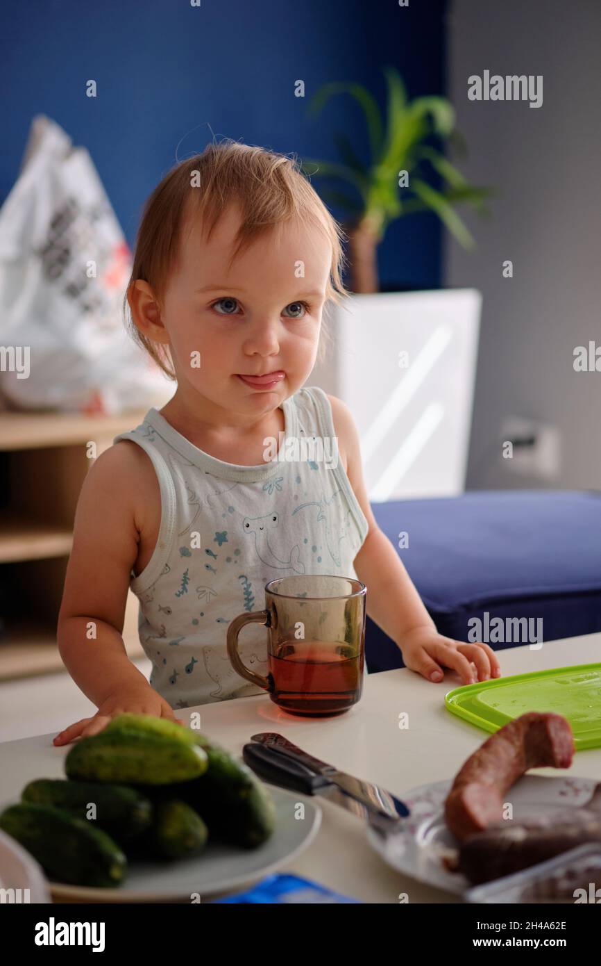 retrato de una niña blanca rubia de pie en la mesa y bebiendo té de un vaso Foto de stock