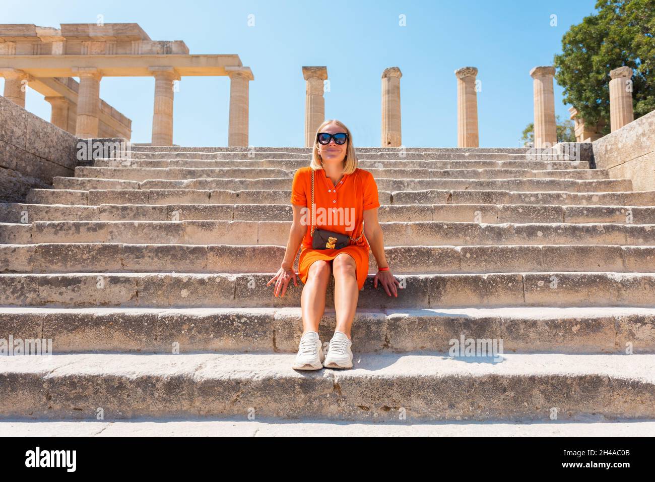 Joven mujer rubia feliz se sienta en los escalones de la Acrópolis en la ciudad de Lindos, Rodas, Grecia Foto de stock