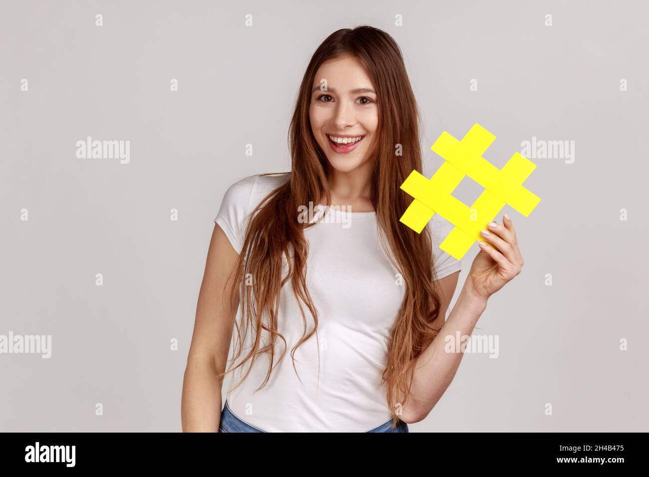Sonriente hermosa mujer sosteniendo el símbolo amarillo hashtag, haciendo popular tema importante, estableciendo tendencias en Internet, usando camiseta blanca. Estudio en interior grabado aislado sobre fondo gris. Foto de stock