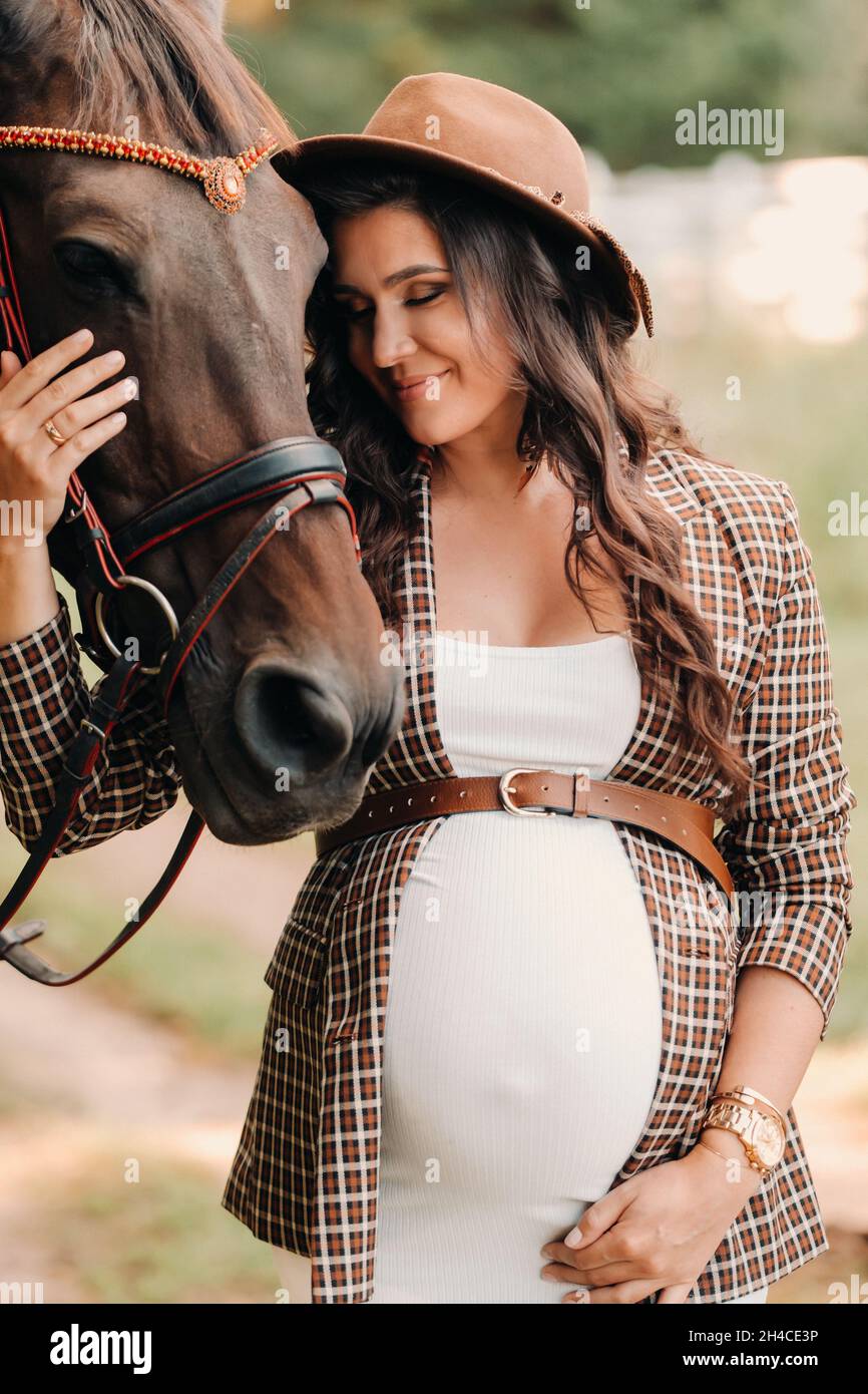Mujer embarazada con un gran vientre en un sombrero junto a los caballos en  el bosque en Natural.una mujer embarazada con estilo en un vestido blanco y  chaqueta marrón con caballos Fotografía