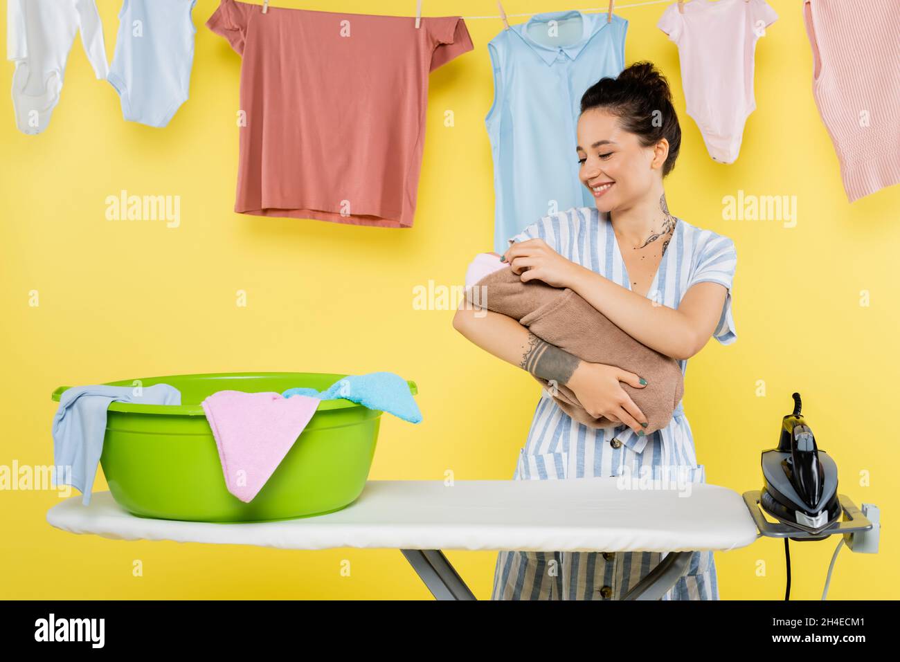mujer sonriente sosteniendo muñeca bebé cerca del recipiente de la ropa en  la tabla de planchar sobre amarillo Fotografía de stock - Alamy