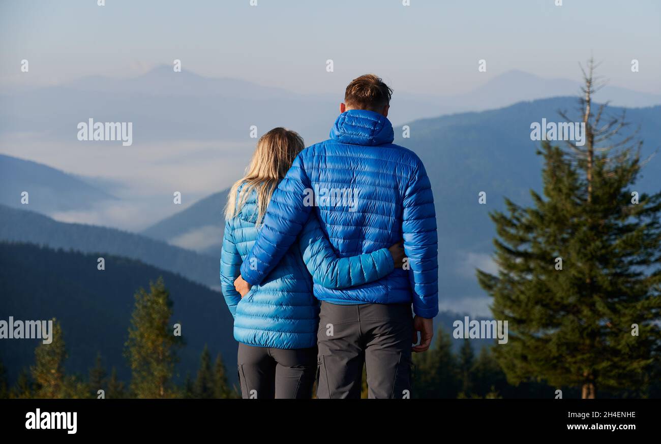 Vista trasera de la pareja joven casada en la misma ropa deportiva disfrutando de la vista de las altas montañas boscosas cubiertas de niebla blanca, abrazándose tiernamente alrededor de la cintura. Foto de stock