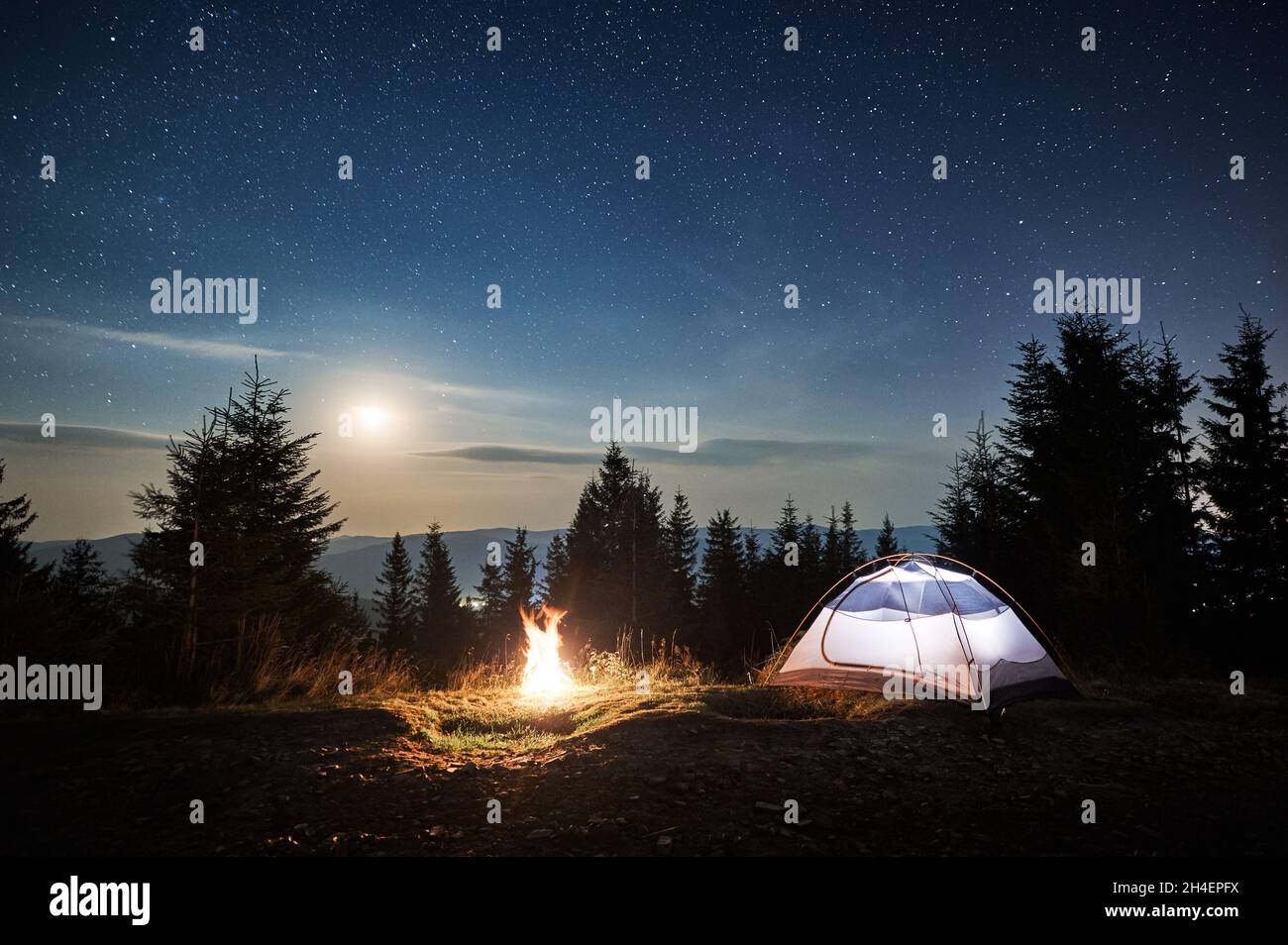 Tienda blanca situada en la colina de montaña cerca de fogatas en el bosque. Luz de luna en el mágico cielo estrellado noche acampando en las montañas. Concepto de viaje, senderismo y acampada nocturna. Foto de stock