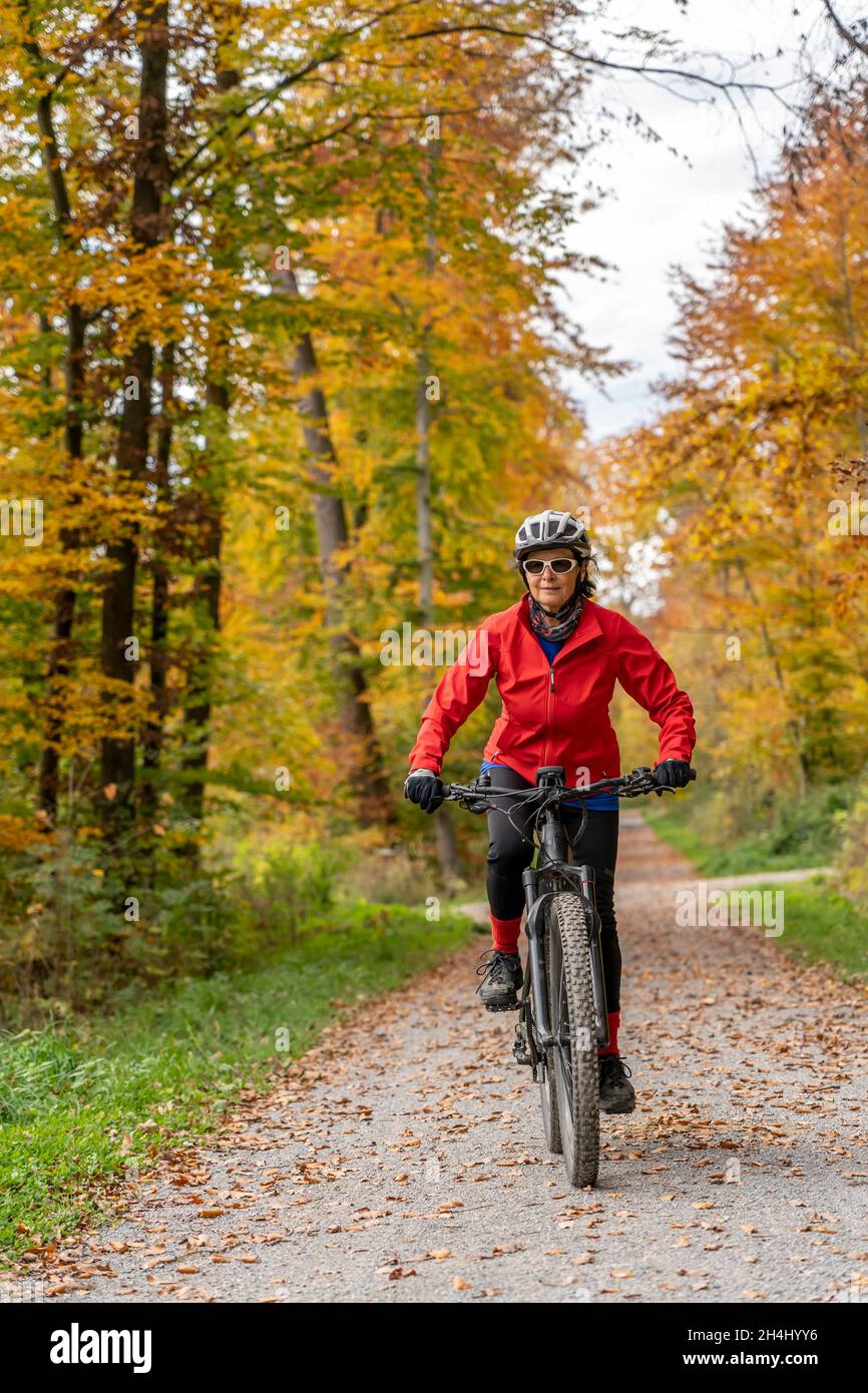 bonita mujer mayor ridin su bicicleta eléctrica en un colorido bosque de otoño con follaje dorado Foto de stock