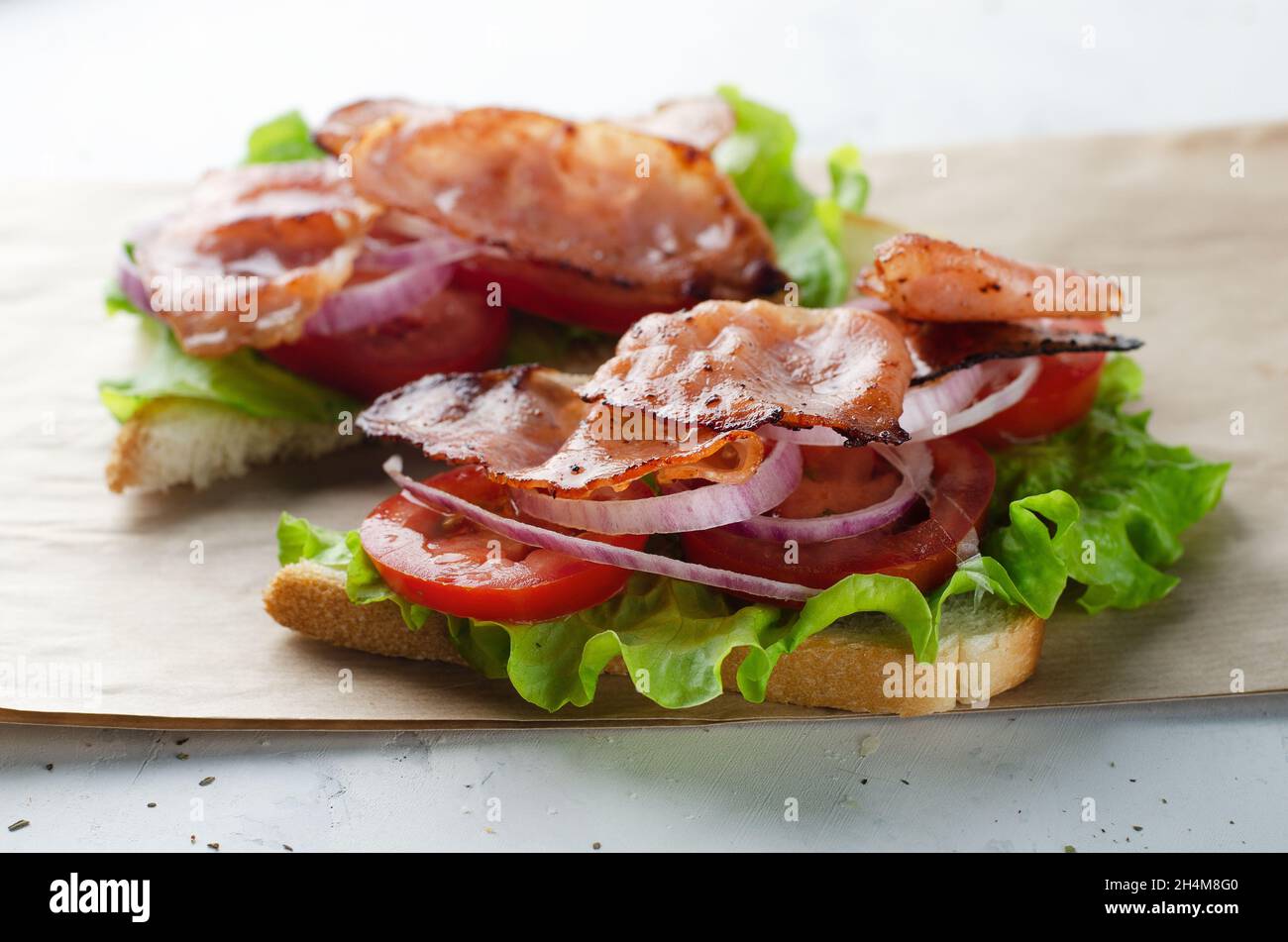 Sándwich de tocino a la parrilla sobre papel con ensalada, tomate y cebolla sobre una mesa blanca Foto de stock