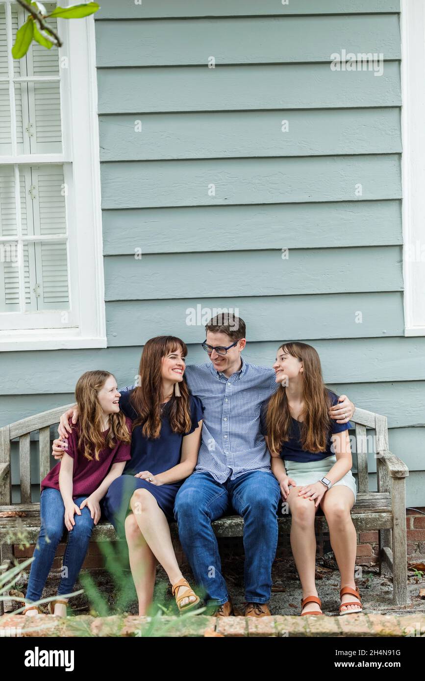 Una familia de cuatro miembros con una madre y un padre y dos hijas sentados en un banco frente a una casa de campo azul Foto de stock