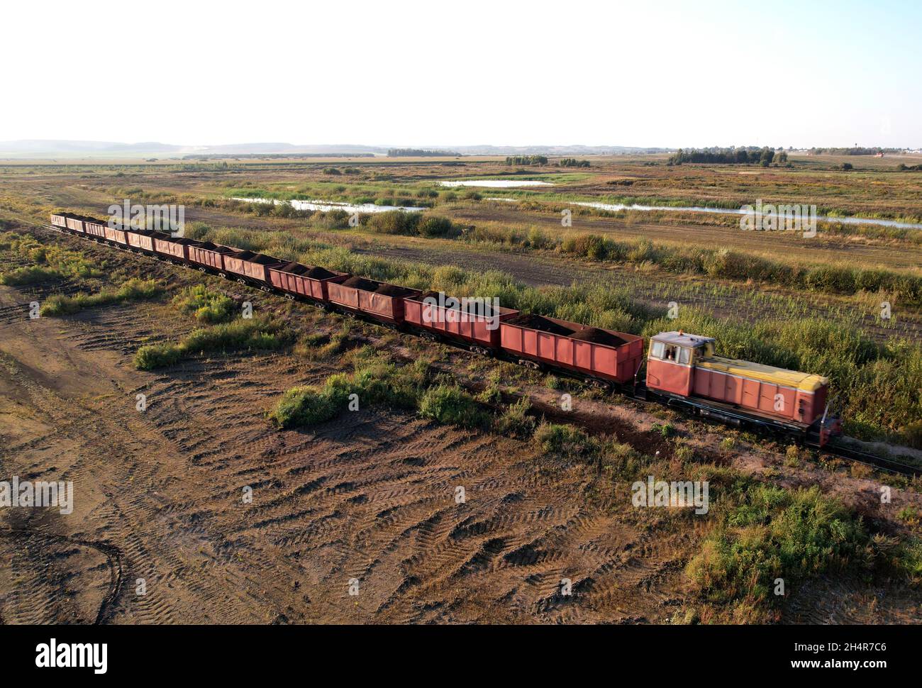 El tren transporta turba en vagones de carga de la extracción de turba.  Vista aérea de la locomotora diesel en el ferrocarril en el paisaje de los  humedales. Vista de drone de