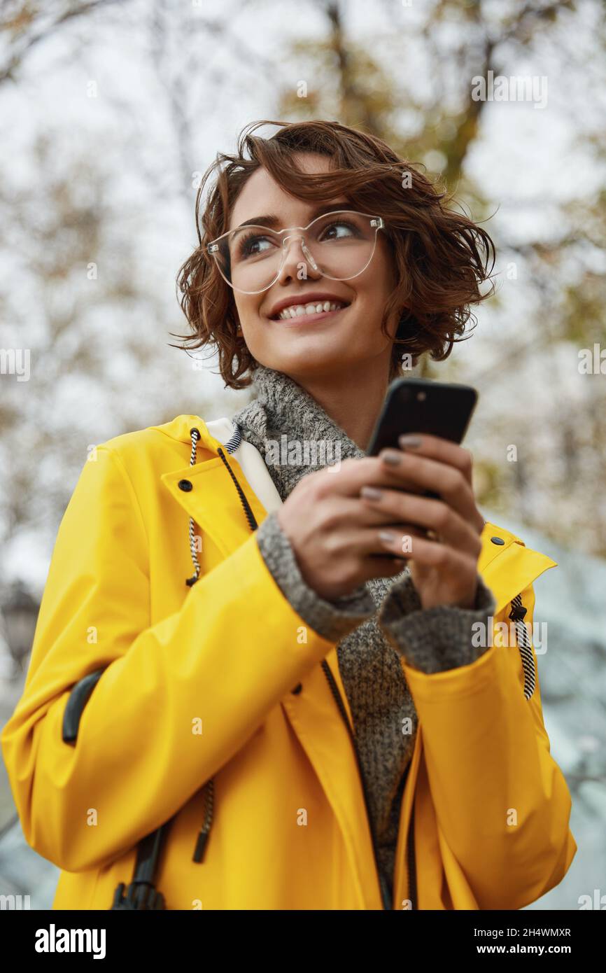 Chica joven con chubasquero amarillo con smartphone en la mano caminando  por la ciudad. Joven hermosa mujer de pelo marrón que lleva traje de lluvia  mientras que va en la stree Fotografía