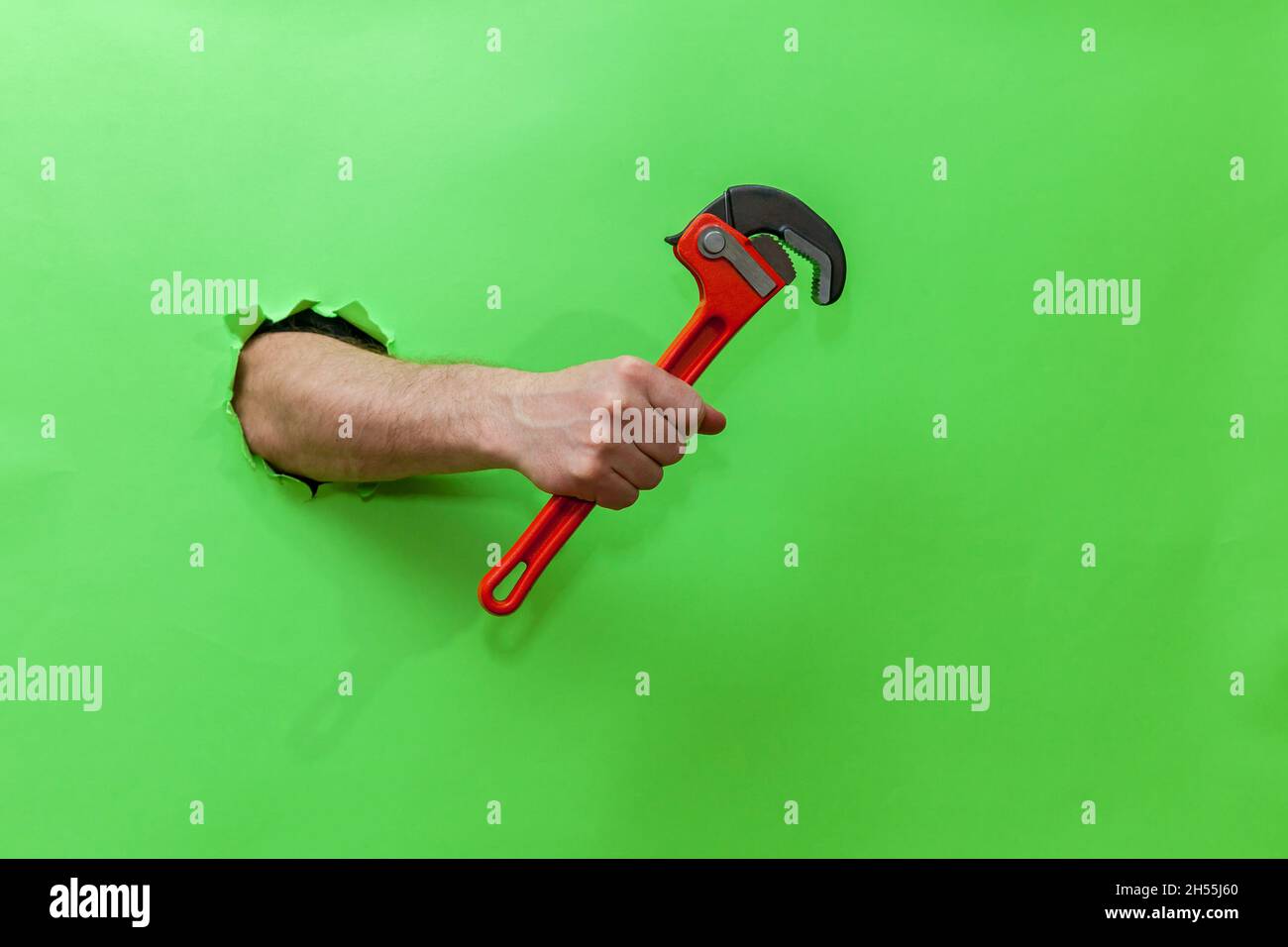La mano macho sostiene una llave roja de la cañería a través de un papel verde rasgado. Mano a través de papel verde rasgado. Foto con lugar para su texto, logotipo y diseño. Representante de concepto Foto de stock
