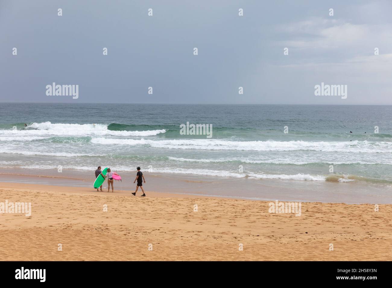Dos surfistas se pararon en la playa de Manly como hombre en negro haciendo ejercicios pasea, Sydney día nublado, Australia Foto de stock