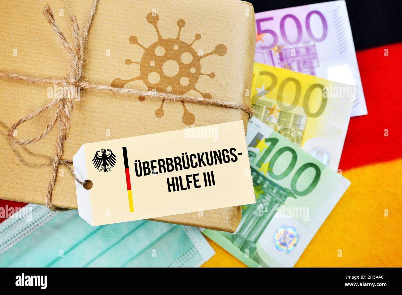FOTOMONTAGE, Paket mit Coronavirus-Symbol auf Deutschlandfahne mit Etikett und der Aufschrift Überbrückungshilfe III Foto de stock