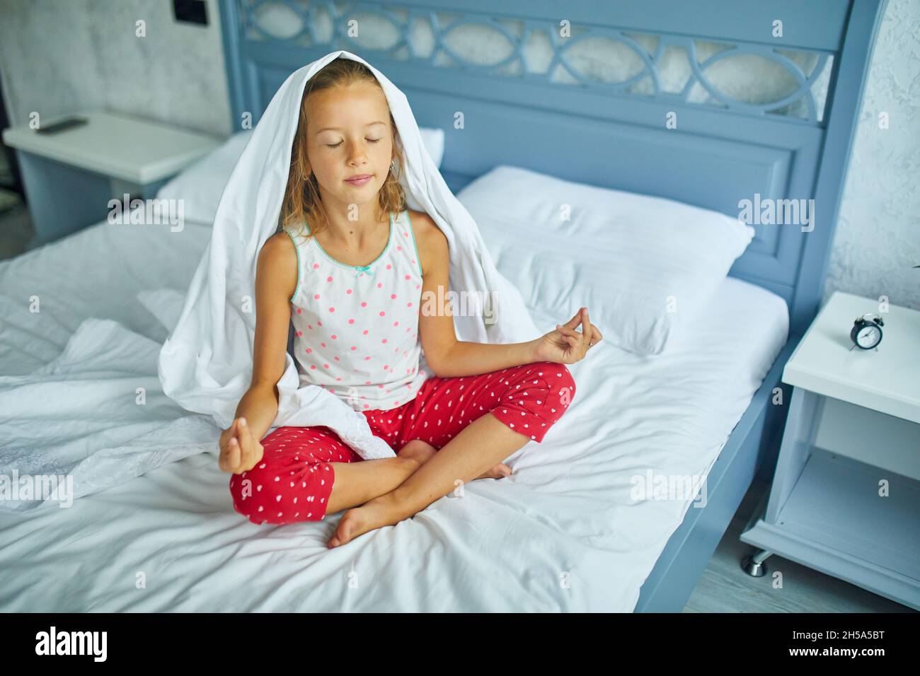 Niña en pijamas sentada en una cama con pose de loto, la niña se despierta  de dormir en una cama grande y acogedora ropa blanca en el hogar, la vida  familiar Fotografía