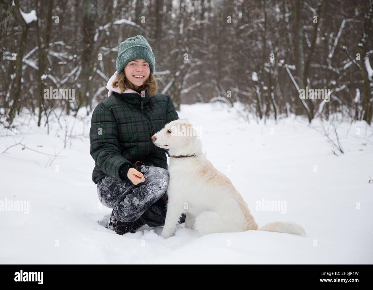 linda niña adolescente sonriente de 16-17 años camina en el bosque nevado con su perro blanco. temporada fría de invierno. Estilo de vida activo, caminando con su querida mascota, jo Foto de stock