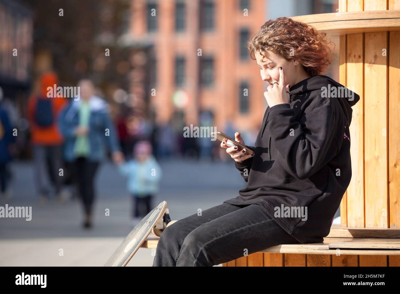 Adolescente con teléfono móvil sentado al aire libre, ropa negra con  capucha y jeans, pelo rizado, copyspace Fotografía de stock - Alamy