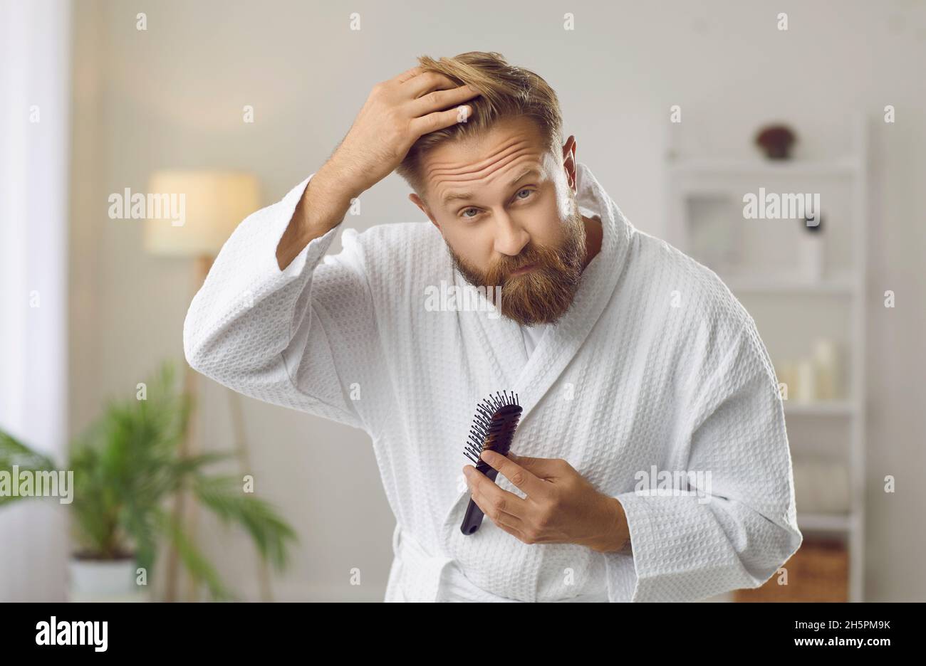 Hombre guapo preocupado con el problema de la pérdida del pelo mirando en espejo en su cuarto de baño Foto de stock