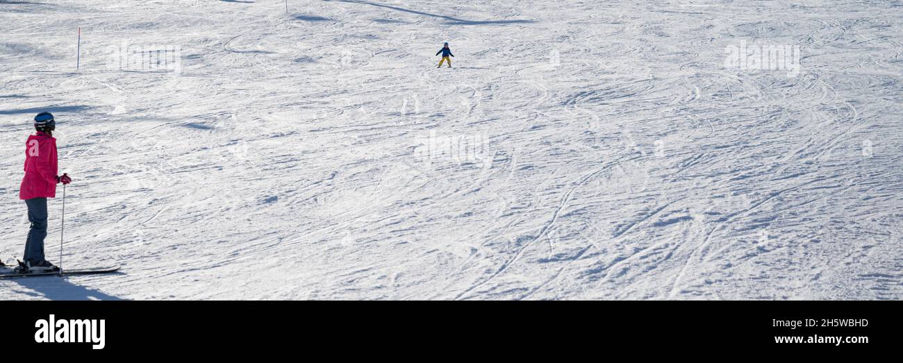 Amplia imagen de una madre en traje de invierno rosa y su hijo esquiando por la ladera blanca nevada. Foto de stock