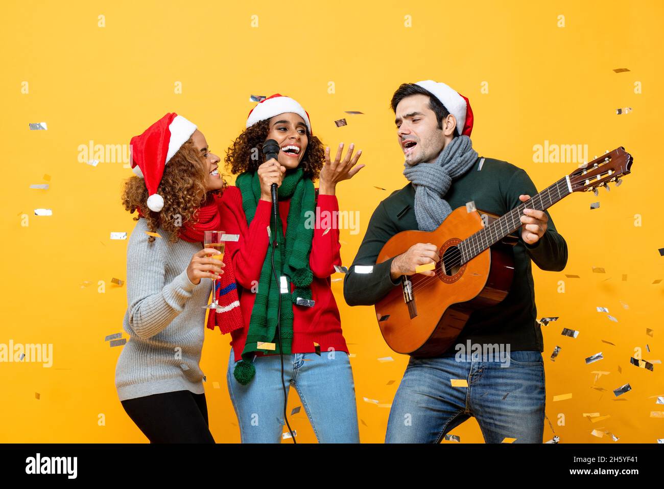 Felices y diversos amigos que tienen fiesta cantando y celebrando la Navidad en fondo de estudio amarillo con confeti Foto de stock