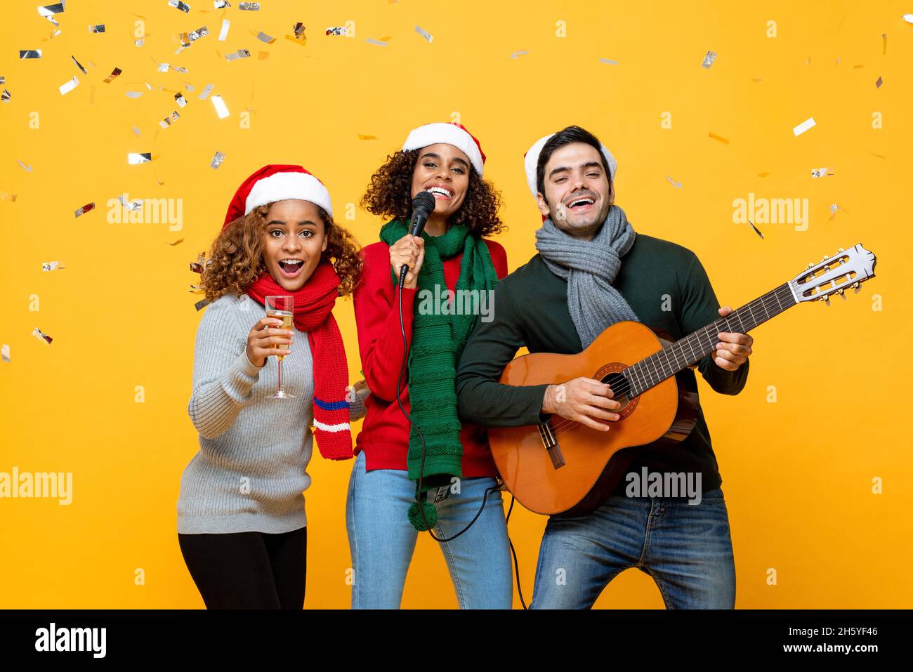 Grupo de amigos cercanos felices y diversos que tienen fiesta cantando y celebrando la Navidad en el fondo amarillo del estudio con confeti Foto de stock