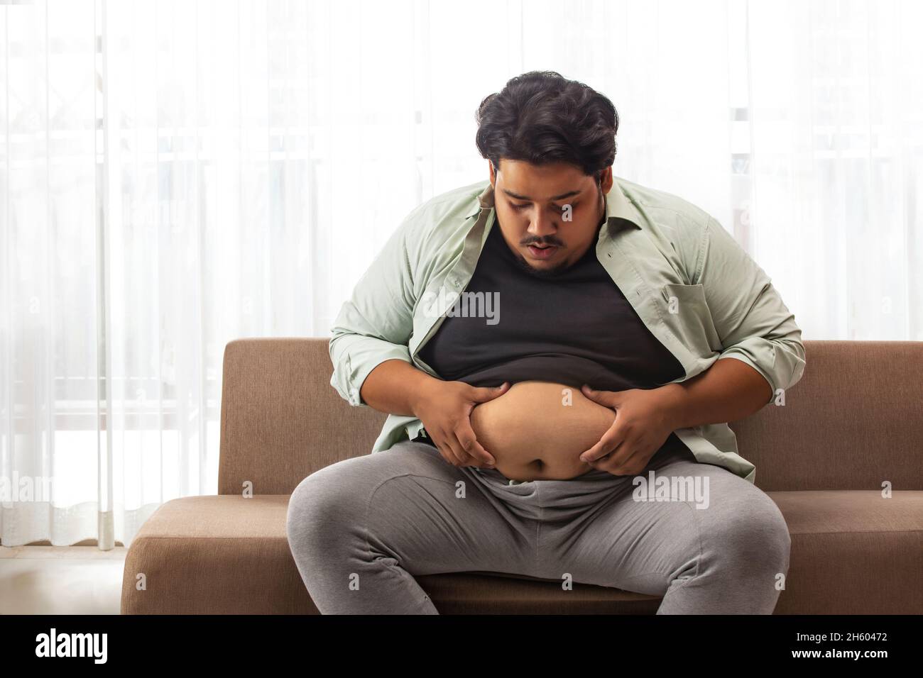 Un hombre gordo sentado en la camilla sosteniendo y mirando la grasa extra del vientre. Foto de stock