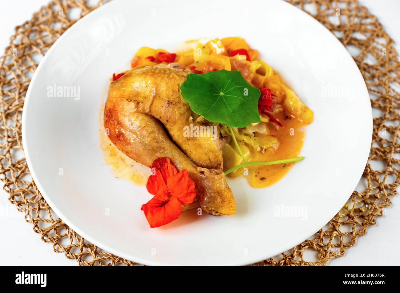 Muslo de pollo horneado sobre pimiento rojo y naranja con fideos, decorado con flores y hojas comestibles en plato blanco y almohadilla de bambú, closeup. Foto de stock