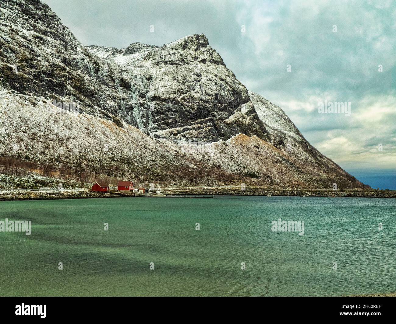 Típicas casas rojas noruegas junto al azul azul azul del mar Ártico en Ersfjord, con cascadas congeladas y nieve en las escarpadas montañas detrás. Foto de stock