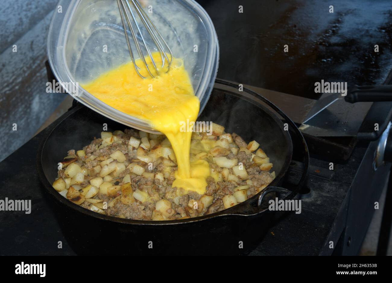 Huevos que se vierten encima de salchichas y patatas fritas en un horno de dtuch; cocina al aire libre de cazuela de desayuno Foto de stock