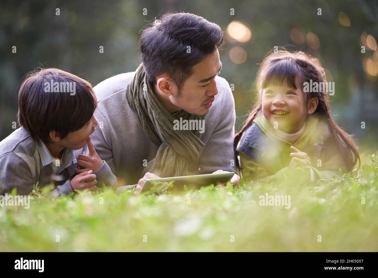 padre asiático acostado en frente en la hierba que cuenta la historia a dos niños feliz y sonriente Foto de stock