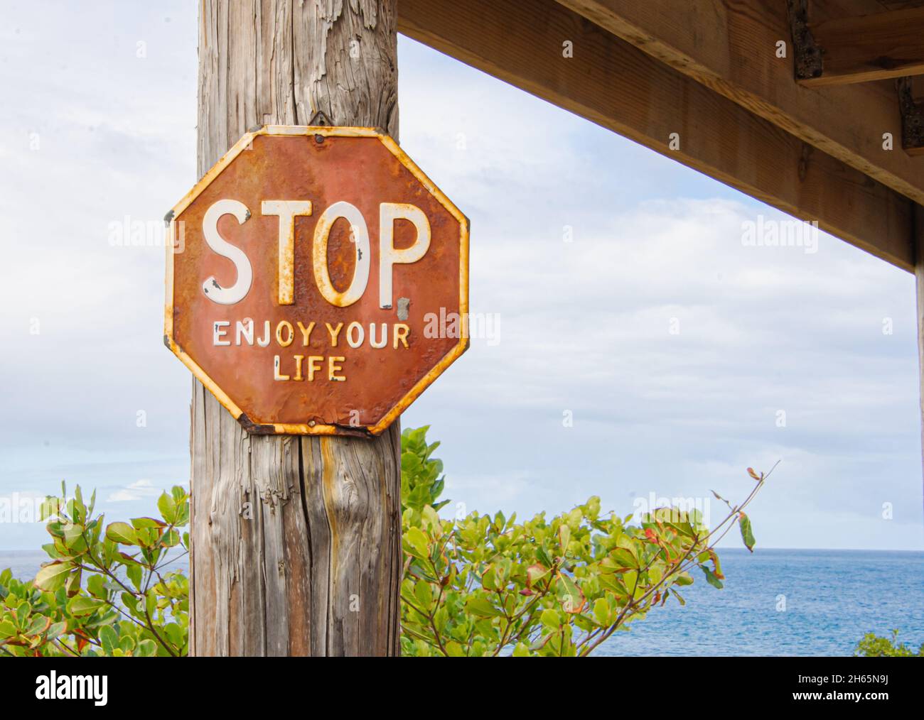 Un cartel octogonal de Stop gastado dice 'STOP Disfruta de tu vida' y está unido a un poste con el azul Mar Caribe en el fondo. Rincón, Puerto Rico. Foto de stock