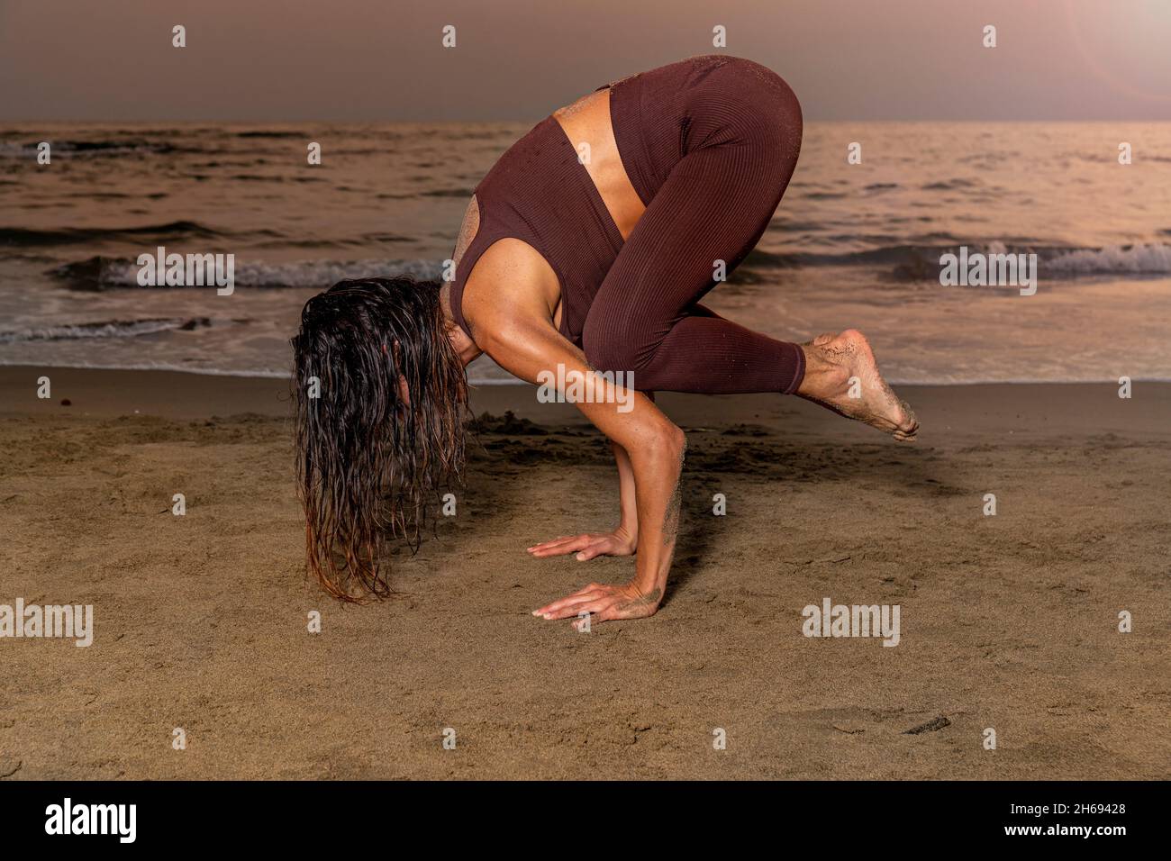 Mujer practicando yoga en la playa al atardecer. Mujer en posición Kakasana, posición de cuervo para concentración en el presente. Foto de stock