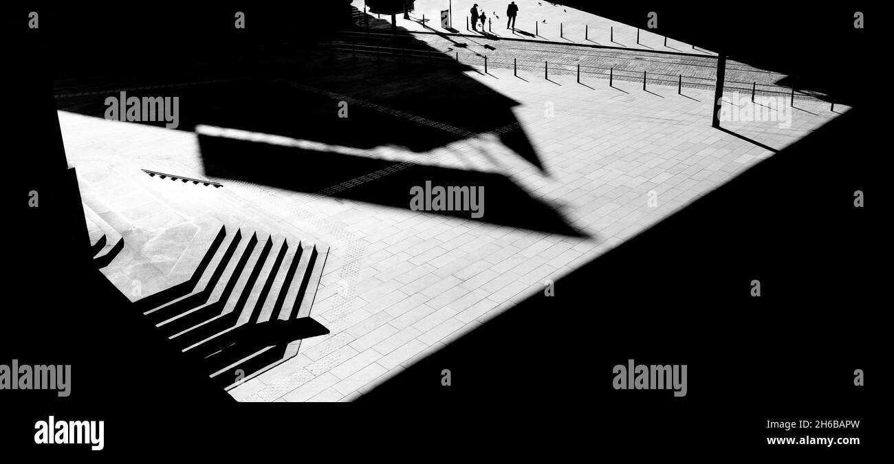 Plaza ancha de la ciudad, espacio público vacío con escaleras en tonos arquitectónicos y luces, y silueta de la gente de pie en la distancia, en negro y w Foto de stock