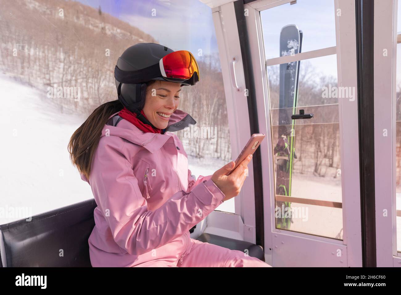 Vacaciones de esquí - Mujer esquiadora usando la aplicación de teléfono en el telesilla. Chica sonriente mirando al smartphone móvil con ropa de esquí, casco y gafas Foto de stock