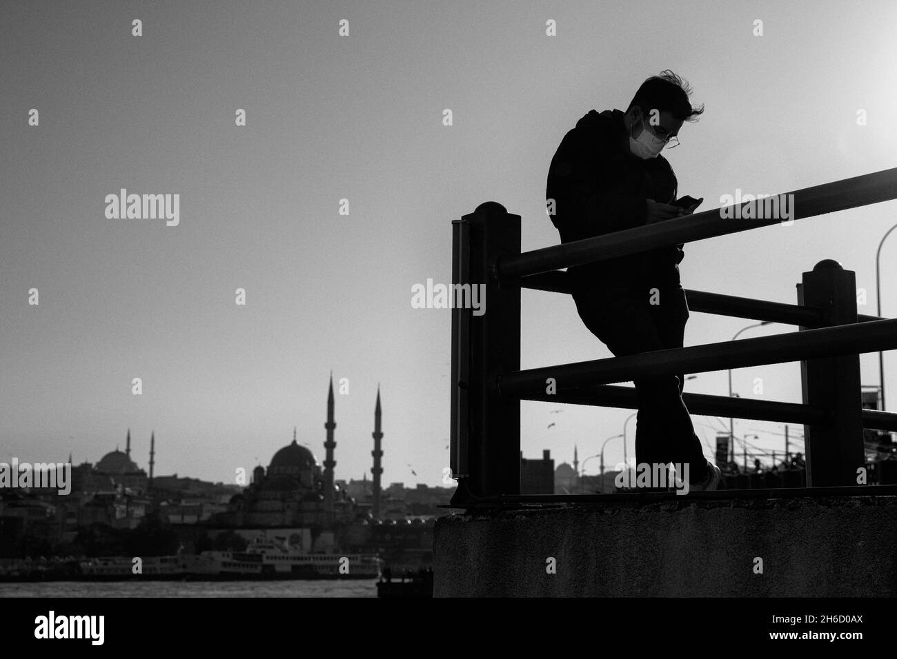 Estambul, Turquía-Nov.12, 2021: Un joven con máscara protectora se encuentra en el puente de Galata y revisa su teléfono móvil. Foto de stock