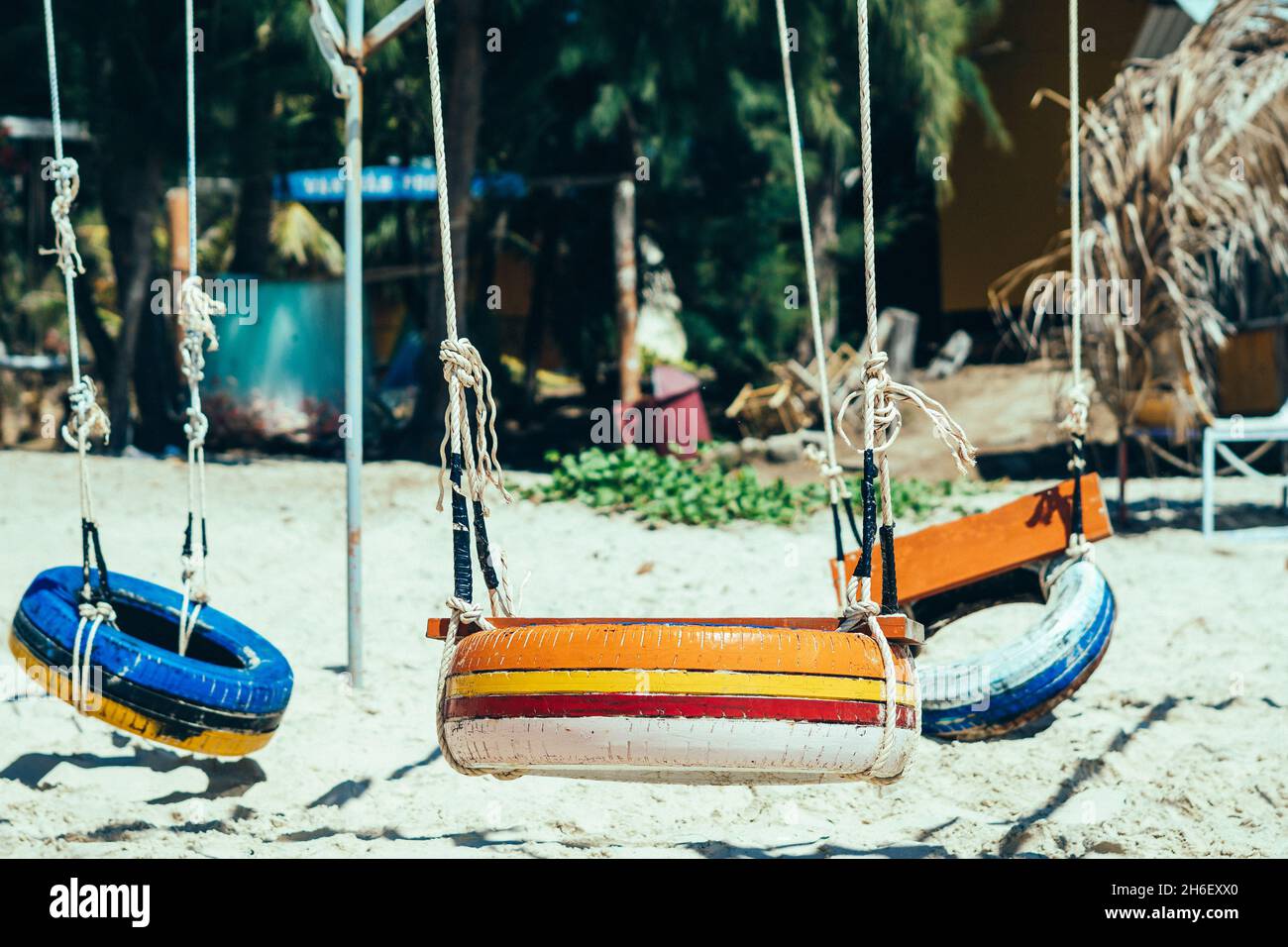 Copiar espacio Hang cuerda swing neumáticos viejos pintura multicolor brillante naranja azul amarillo. Naturaleza arena playa palmeras fondo. Día sol verano simple alegría diversión Foto de stock