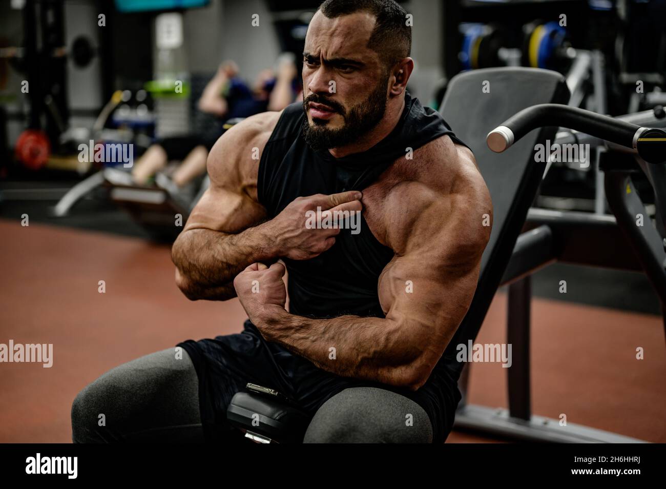 hombre fuerte barbudo culturista mirando su músculo de hombro y brazo sentado en un gimnasio deportivo Foto de stock
