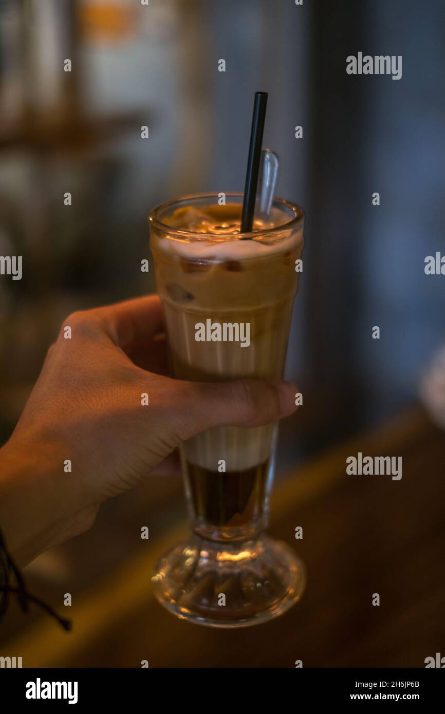 Foto de primer plano de una taza de cristal con leche helada en una cafetería. Mano sosteniendo el vaso con capuchino caramelo helado con la paja en el restaurante. Foto de stock