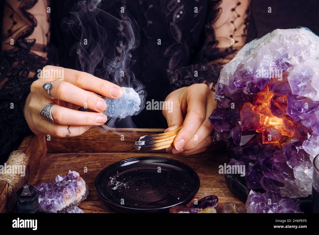 Mujer glamorosa en negro vestido limpiando cristales piedras preciosas por el palo de madera de Palo Santo. Elimine la energía negativa. Foto de stock