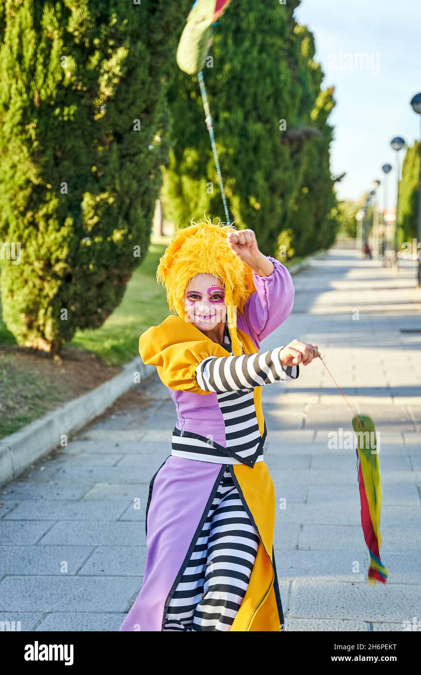 Feliz payaso femenino con rostro pintado con divertido traje y peluca amarilla, sonriendo y caminando por el camino en el parque con coloridos poi en las manos durante el desfile Foto de stock